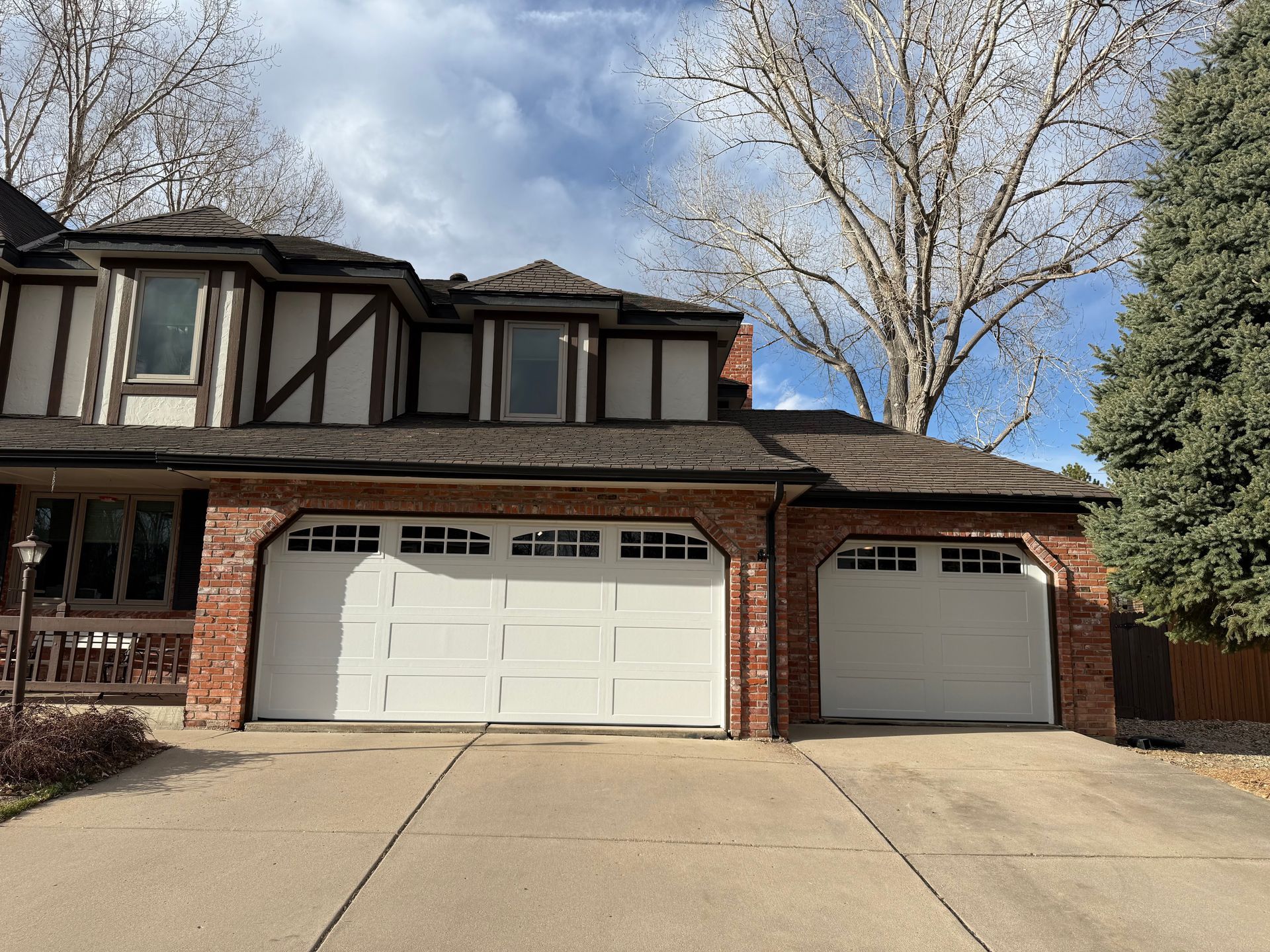 A two-story Tudor-style home with a two-car garage, a single-car garage, and cream stucco walls with dark wood trim.