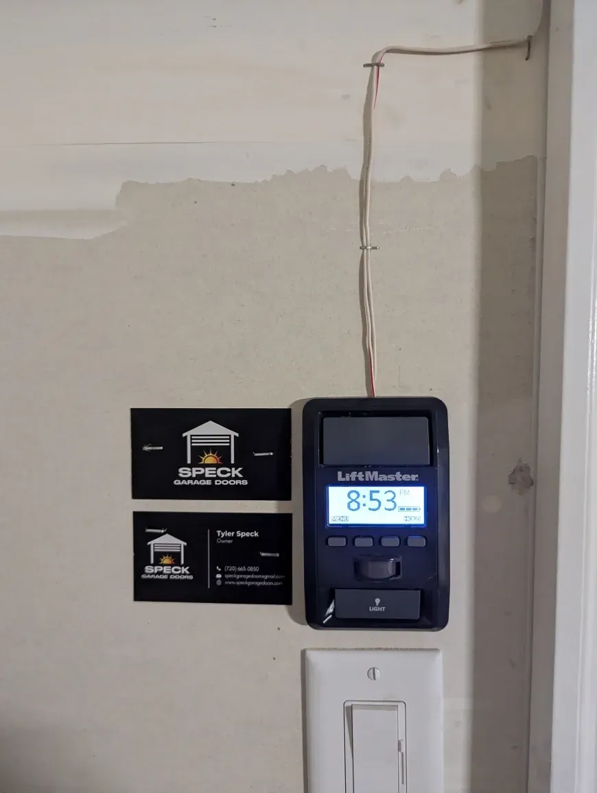 A LiftMaster garage door opener wall button with a digital display showing 8:53 next to two business cards on a wall.