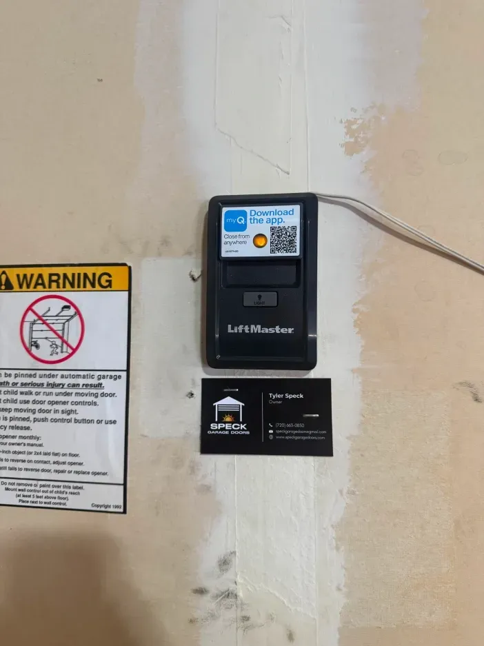 A garage door controller mounted on a wall next to a warning sign and a business card for Open Sesame Garage Doors.
