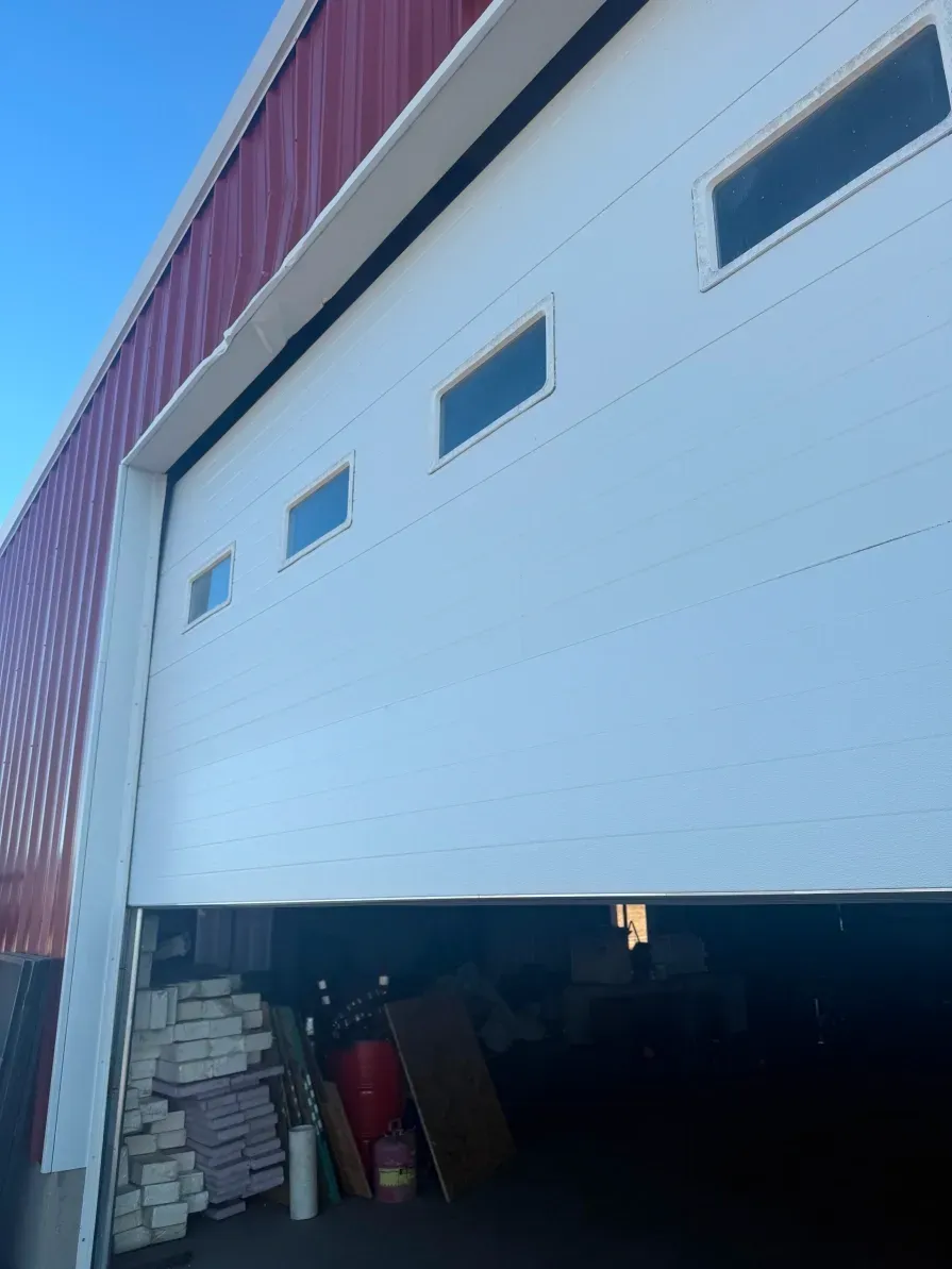 A partially open white industrial garage door with four windows, attached to a red metal-sided building.