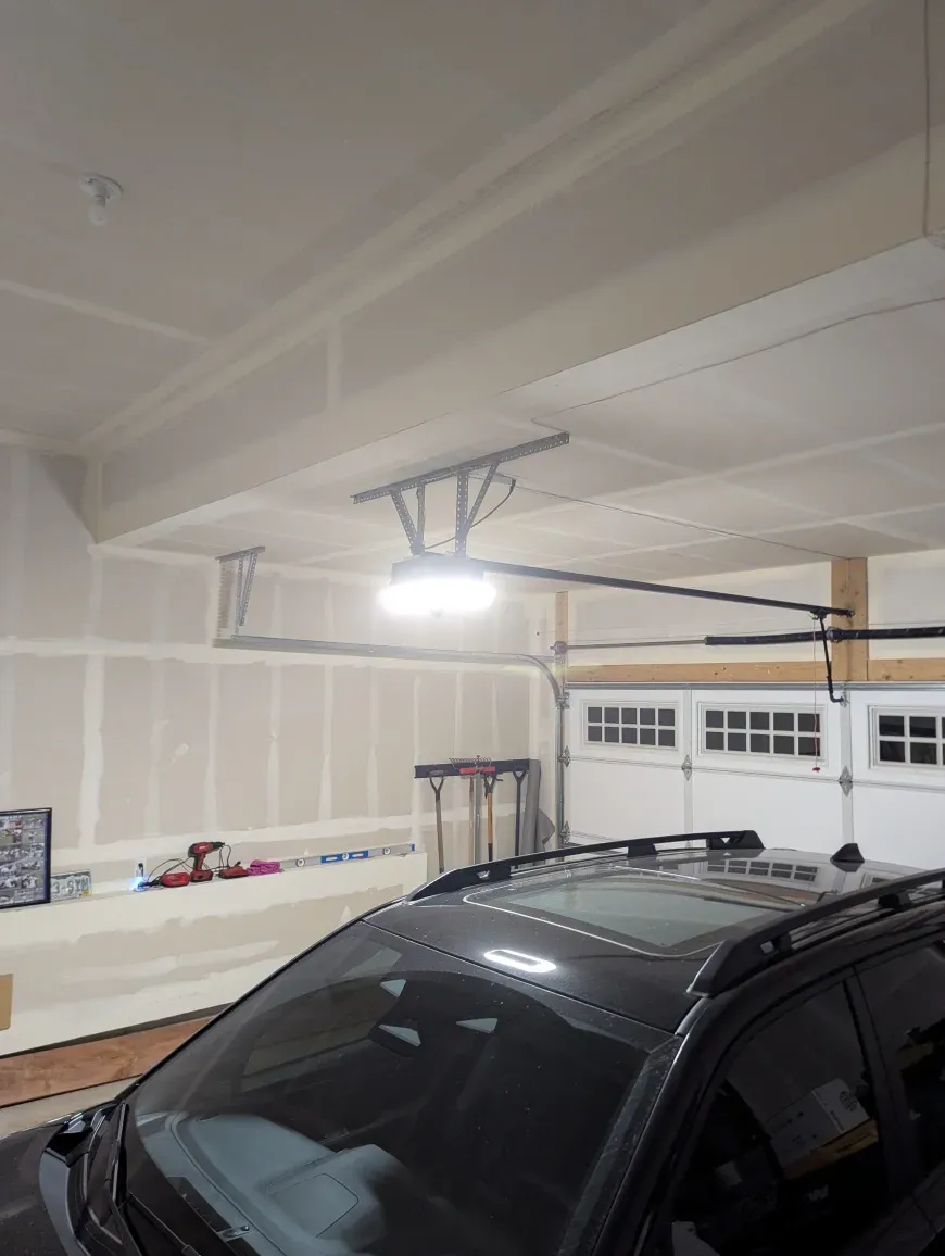 An interior view of a garage with a lit ceiling-mounted garage door opener above a parked black car.