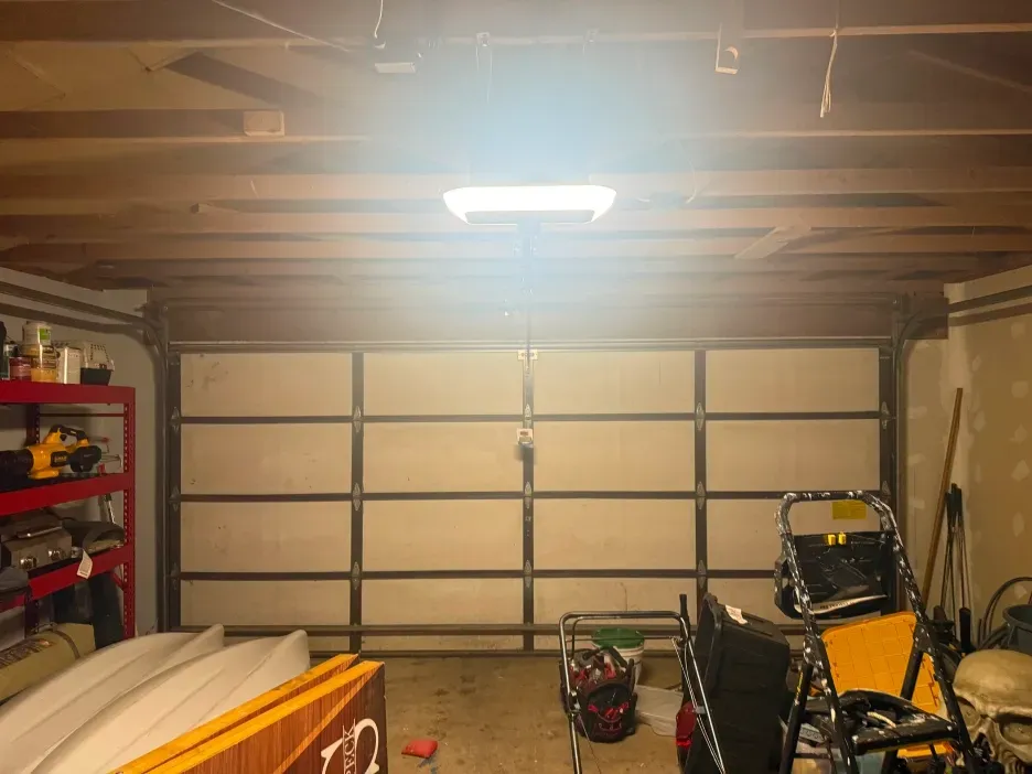 A garage interior with a large sectional door, storage shelving, kayaks, and a ladder under a bright ceiling light.