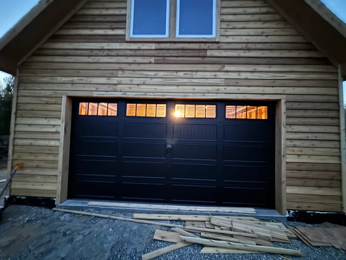 A detached wooden garage with a black, windowed door and upper-level windows under construction on a gravel lot.