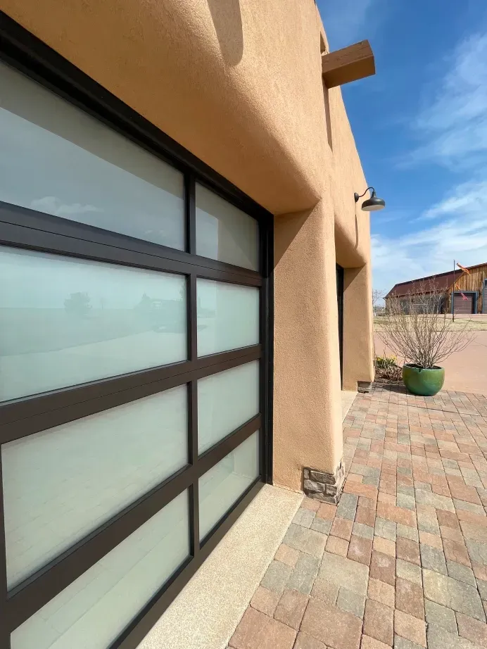 Modern garage door with frosted glass panes set in a dark frame on a tan stucco house with a stone-paved driveway.