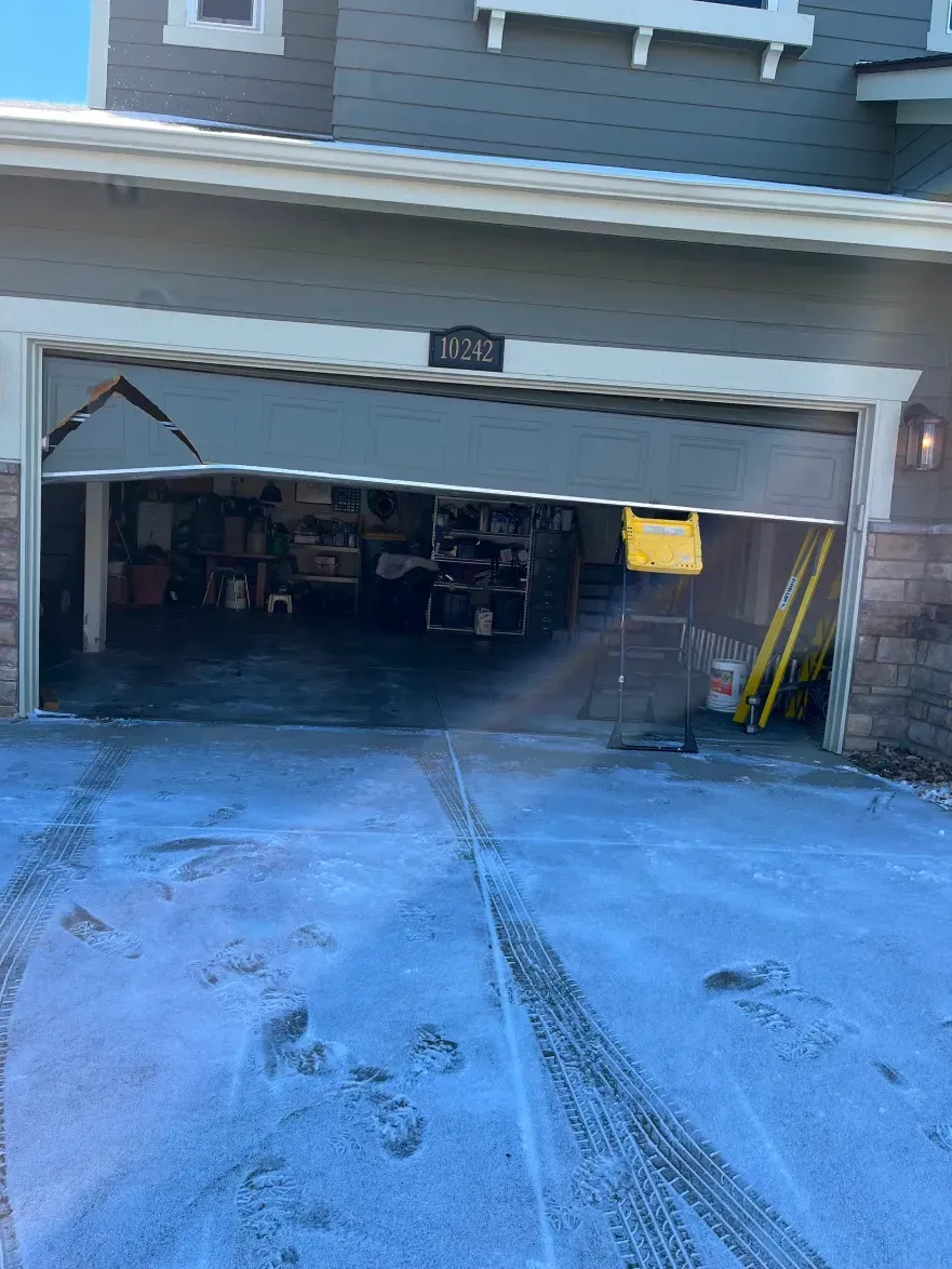 A garage door hangs crookedly from its tracks on a house with a snowy, tire-tracked driveway.
