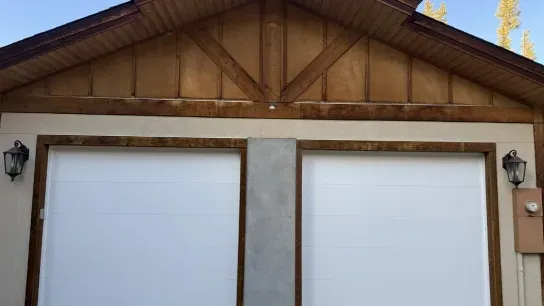 A two-car garage with white doors, framed in dark wood, with wooden gable siding above and wall-mounted exterior lamps.