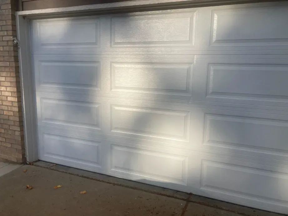 A white, paneled garage door viewed from an angle against a brick wall and concrete driveway.