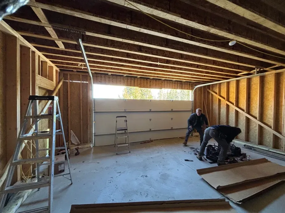 Two people install a garage door in an unfinished wooden workshop with a metal ladder nearby.