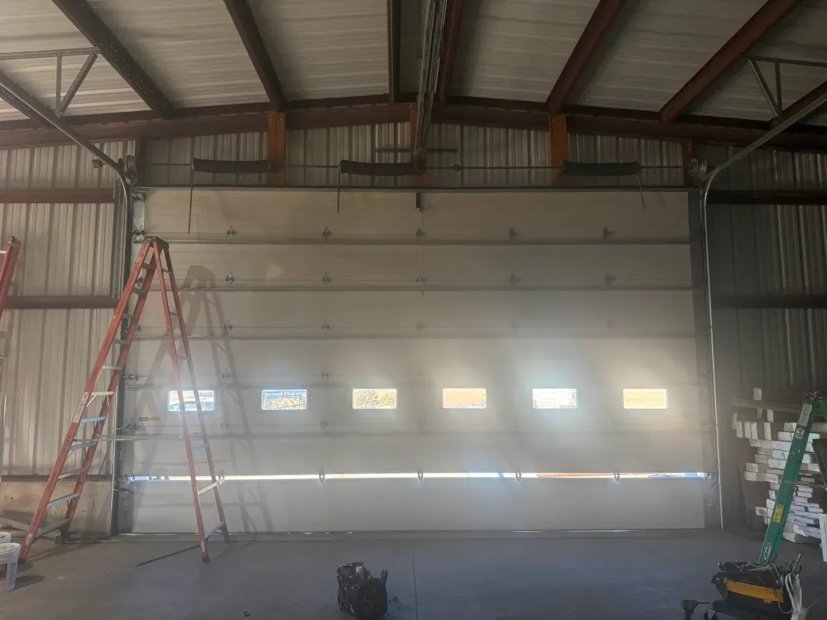 A large white commercial sectional garage door with a row of windows in an industrial warehouse setting with two ladders.