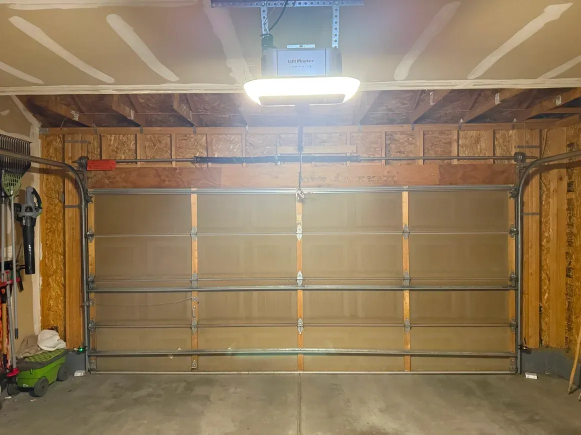 A closed, tan, horizontal-paneled residential garage door with a central overhead opener and exposed wooden wall framing.