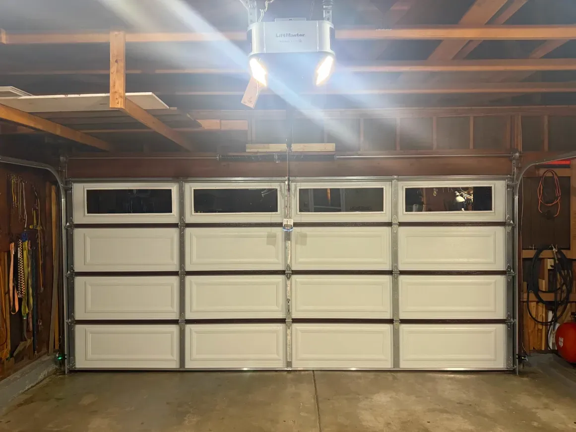 White sectional garage door with four top window panels, viewed from inside a wooden garage with a ceiling-mounted opener.