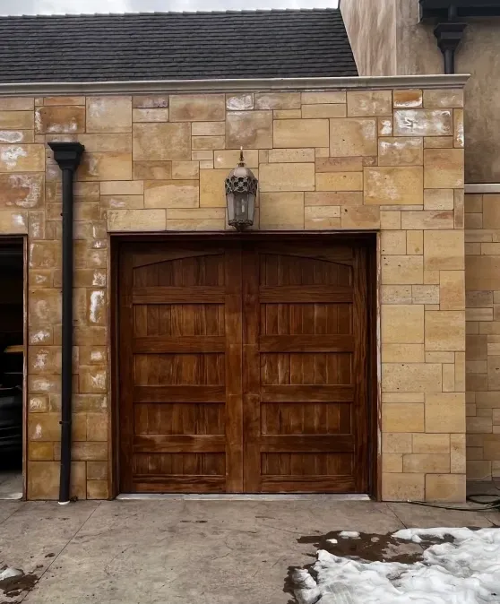 A brown wooden carriage-style garage door set in a tan stone wall with a vintage lantern mounted above it.