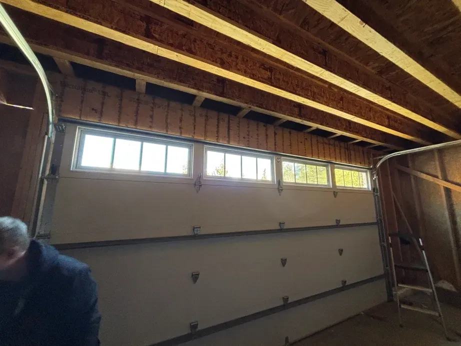 A white garage door with rectangular windows installed in a wooden garage frame with exposed ceiling joists.