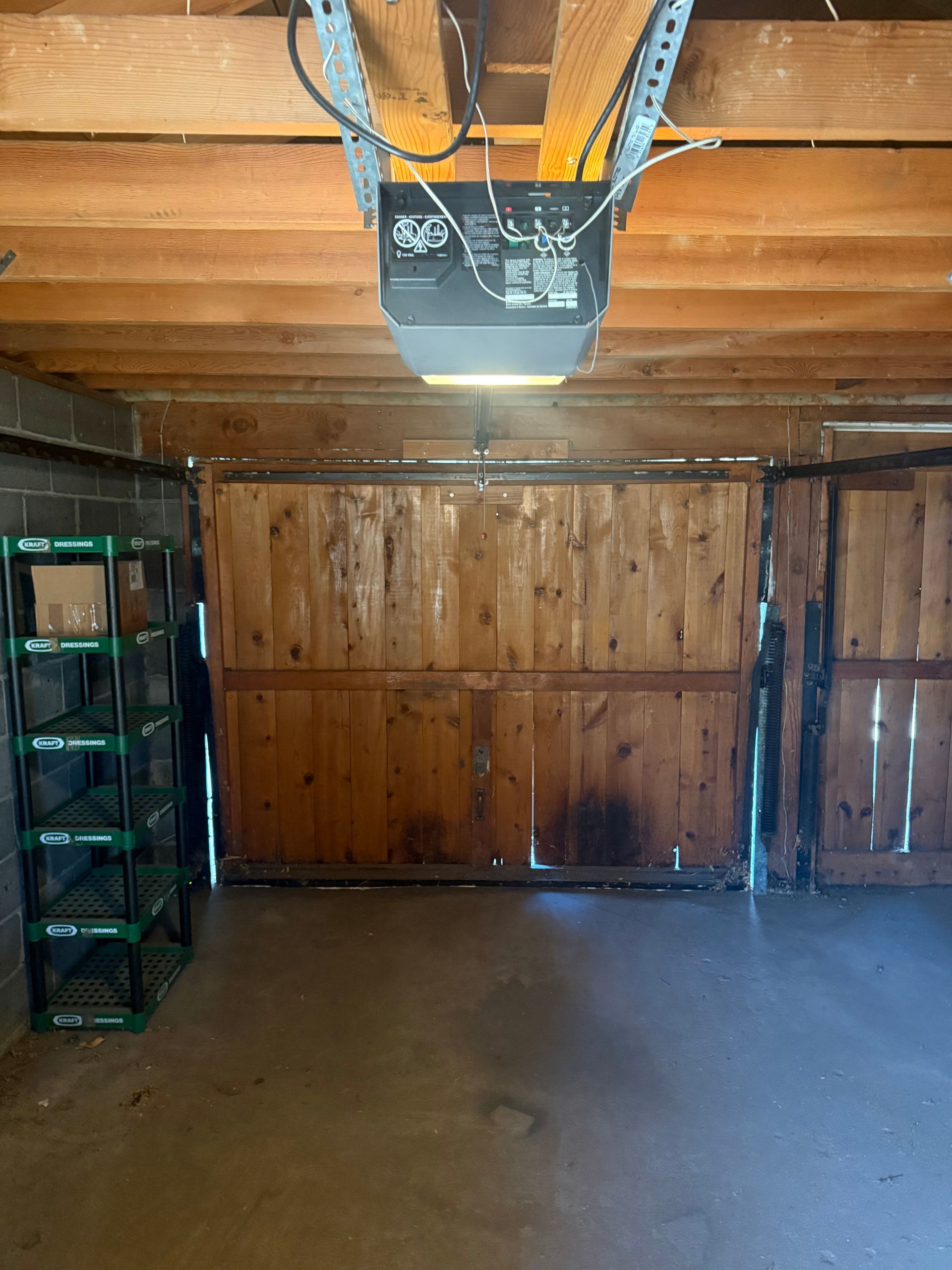 A wooden garage door with a central electric opener mounted to exposed ceiling joists and a plastic shelving unit.