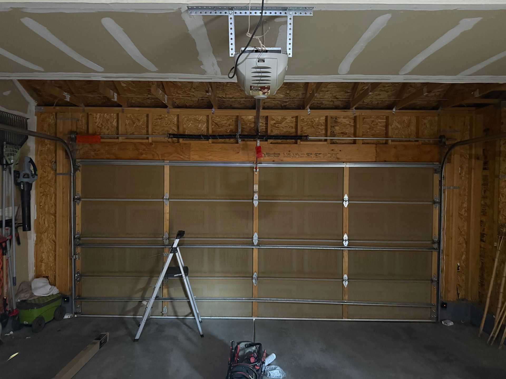 A garage interior showing a sectional door with a mounted automatic opener, a step ladder, and a tool bag on the floor.