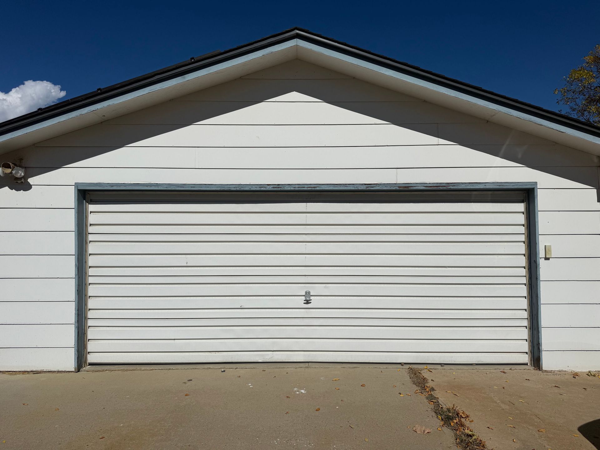 White garage with horizontal siding and a large, closed white garage door under a peaked roof against a blue sky.