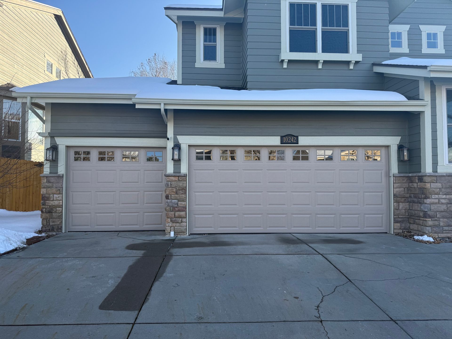 A gray two-story house with a three-car garage, stone accents on the corners, and a light dusting of snow on the roof.