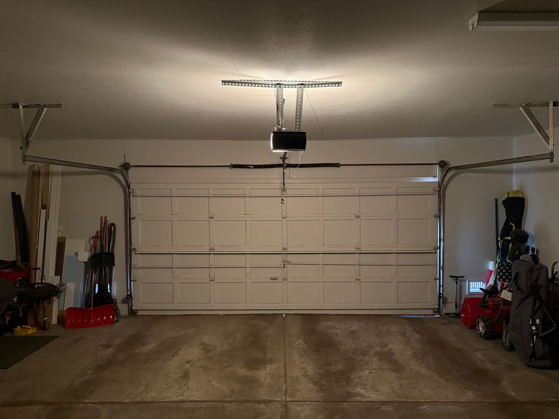 Interior view of a residential garage with a white sectional door, mechanical opener, and storage items on both sides.
