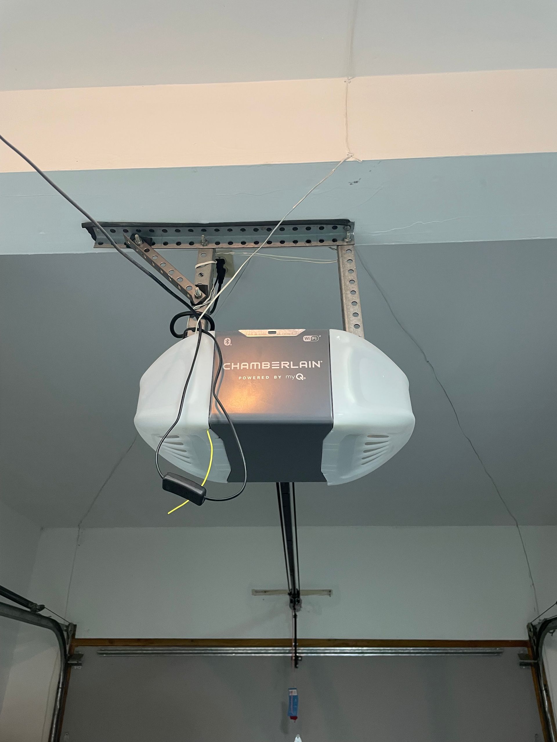 A gray and white garage door opener mounted to the ceiling with exposed wires hanging near the motor unit.