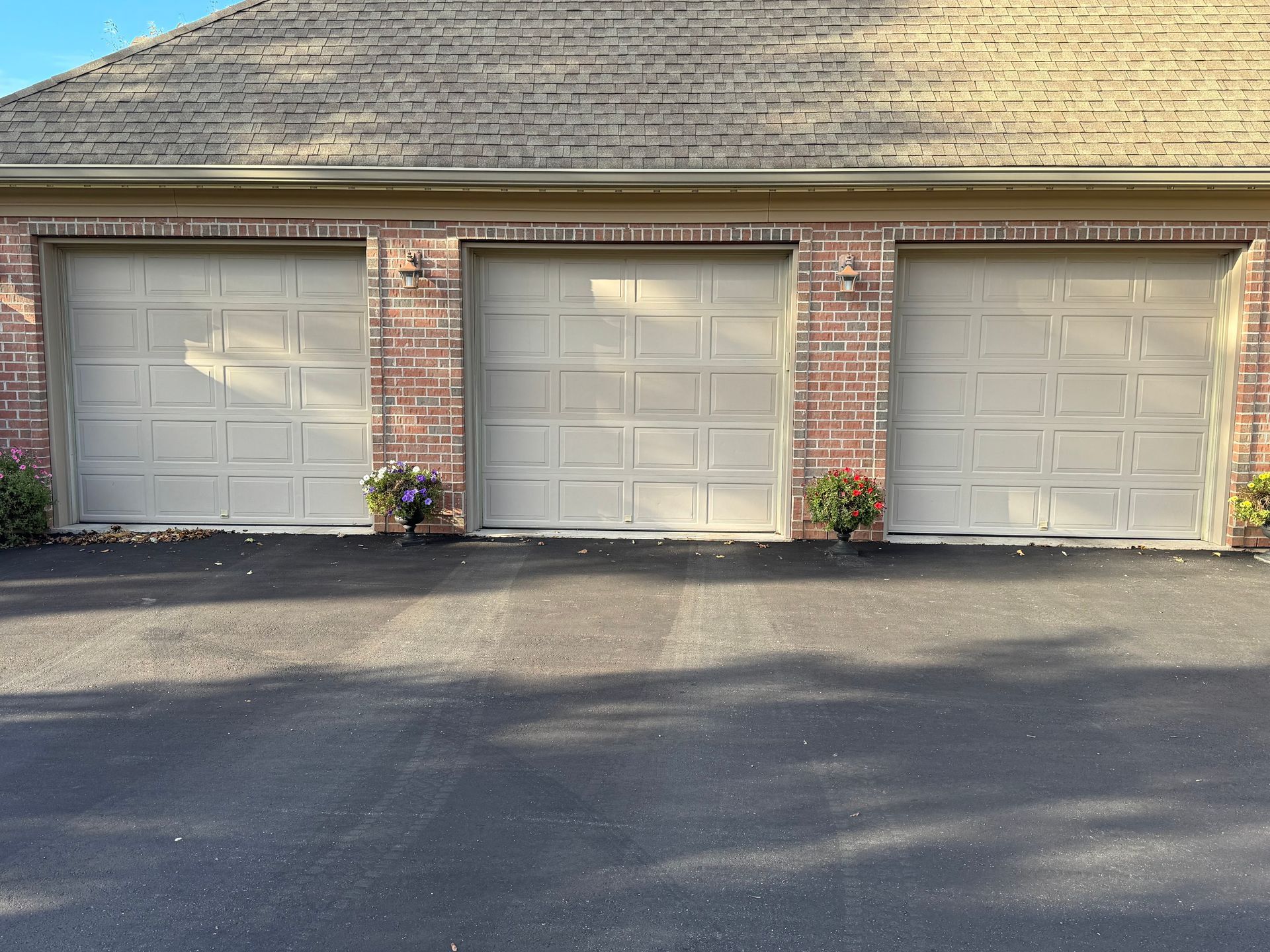 A three-car garage with beige paneled doors and brick columns, featuring potted plants between the bays.