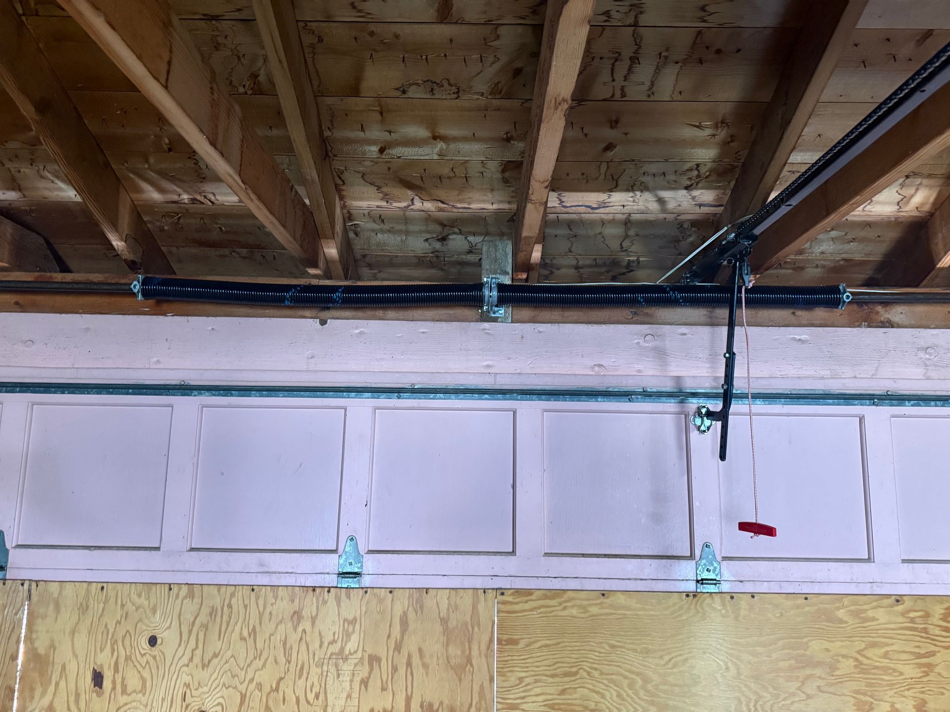 A garage door torsion spring system mounted on a wooden header above a pink-painted garage door.