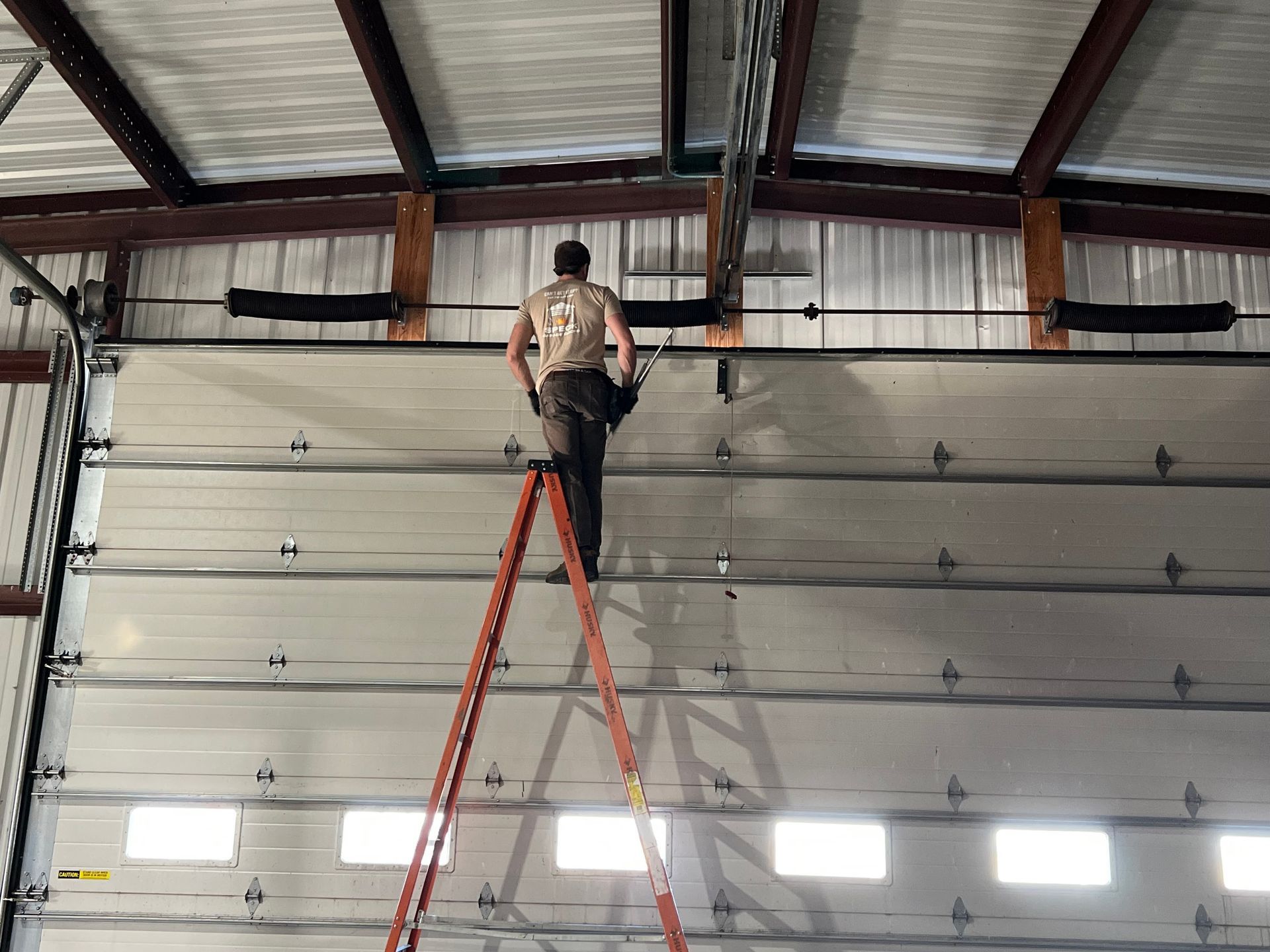 A person stands on a tall, orange ladder working on the top tracks of a large, white commercial garage door.