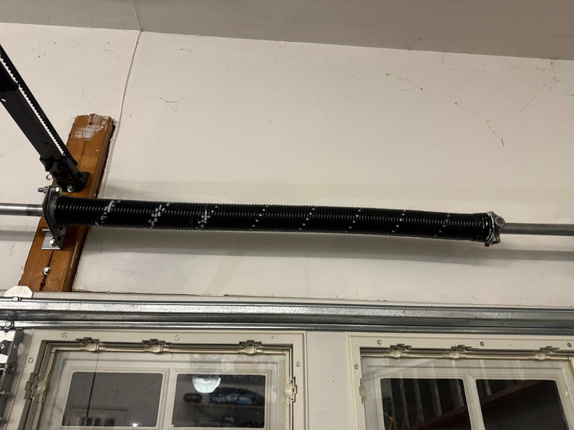 A black garage door torsion spring mounted horizontally on a metal bar above a garage door.