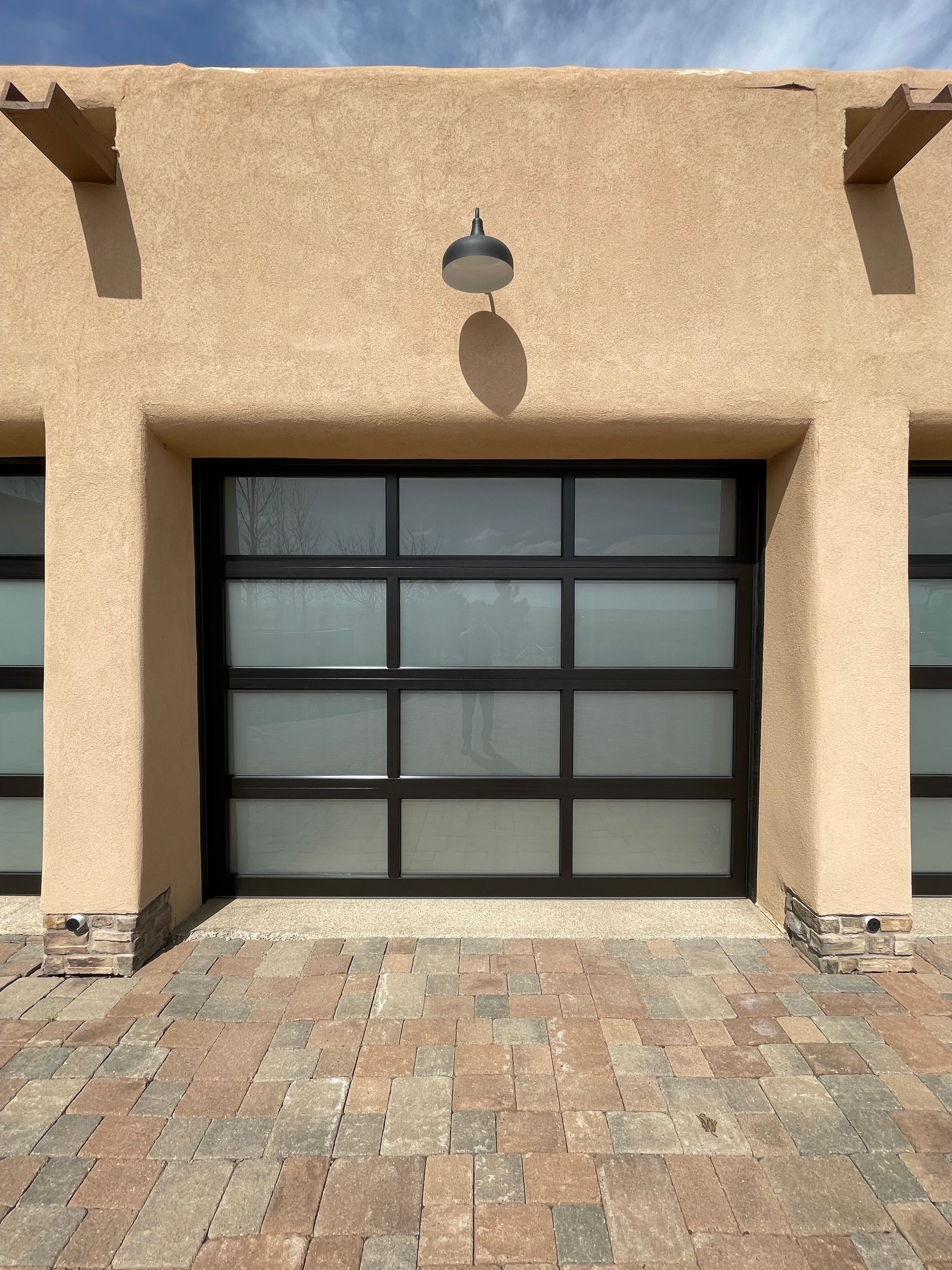 A modern garage door with frosted glass panels and a dark frame set in a textured stucco building with a brick driveway.