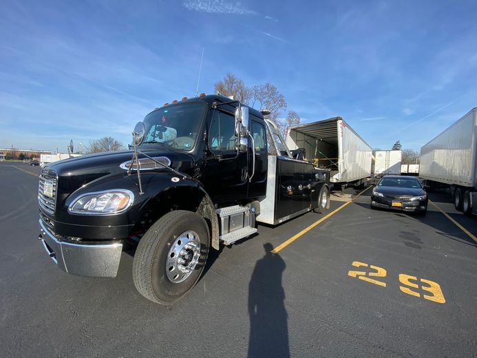 Black tow truck parked, hitched to a trailer, in a sunny parking lot.