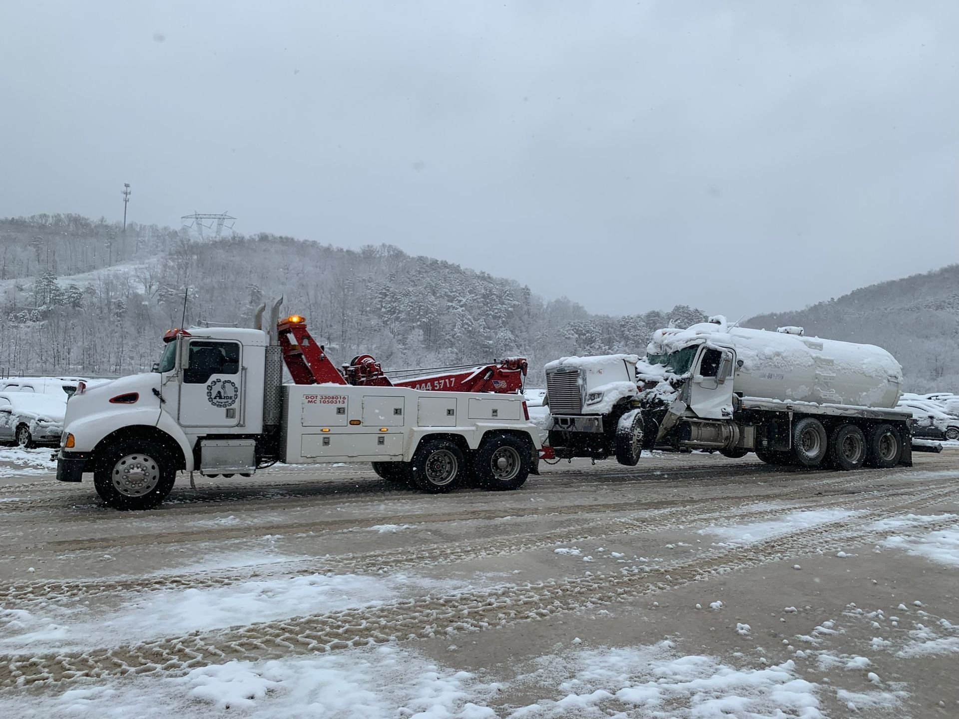 Tow truck towing a large tanker truck in a snowy parking lot.