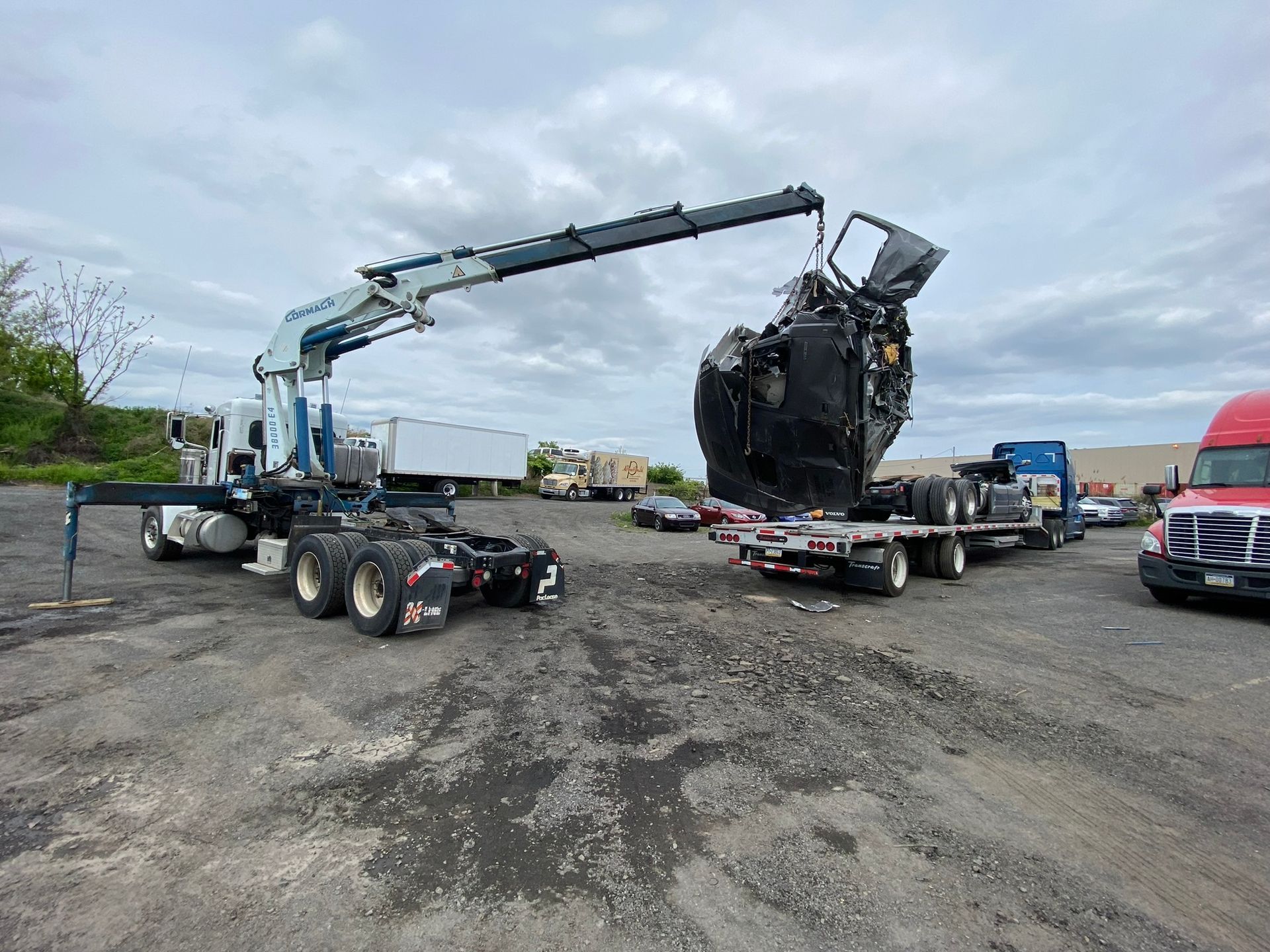 Crane lifting a crushed semi-truck cab onto a flatbed trailer in a salvage yard.