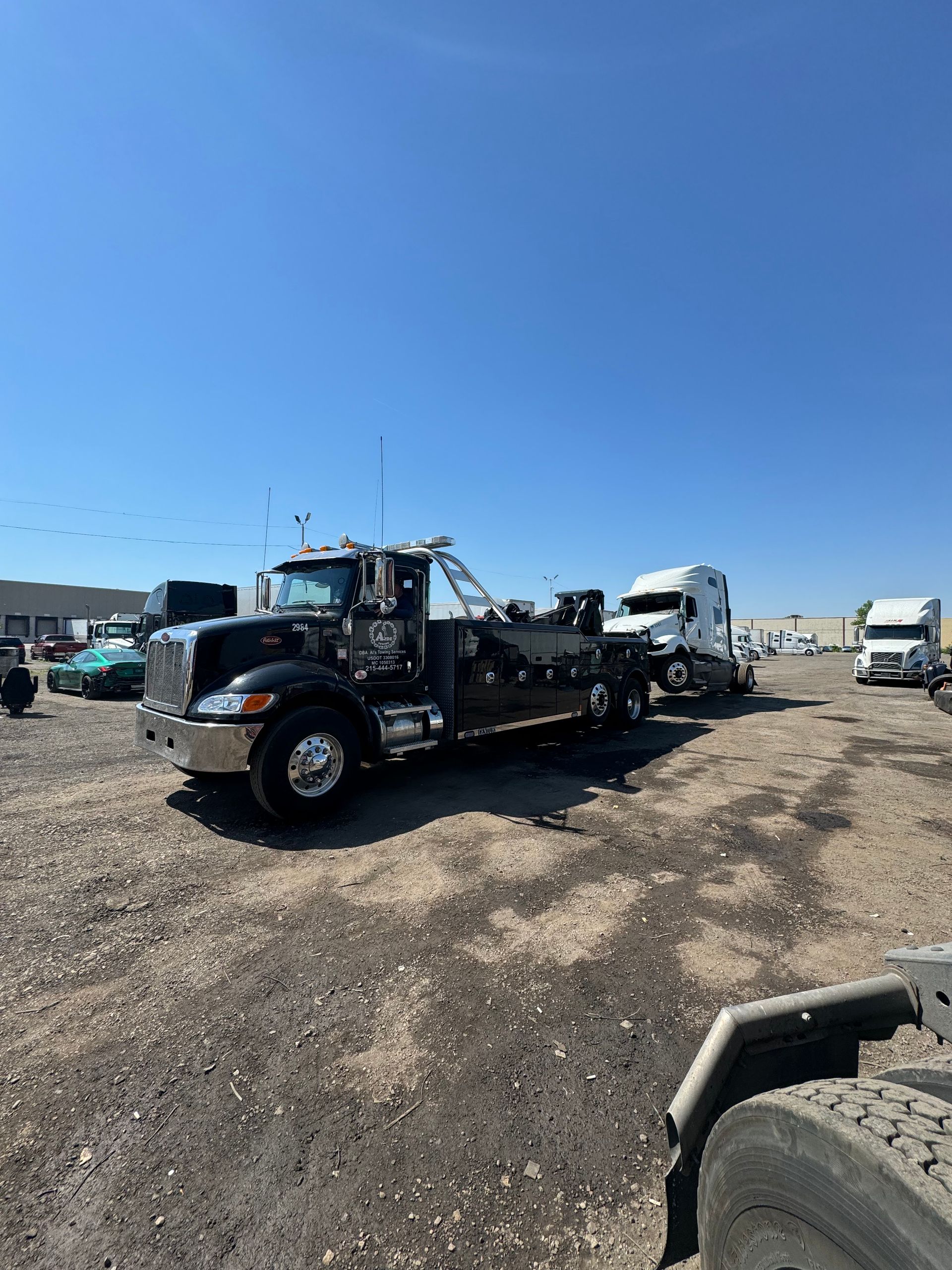Black tow truck towing a white semi-truck on a gravel lot under a blue sky.