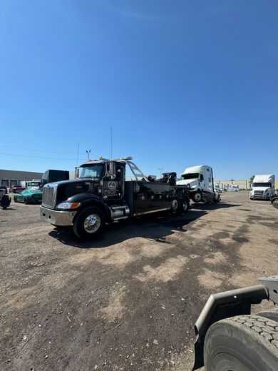 Black tow truck towing a white semi-truck on a gravel lot under a blue sky.