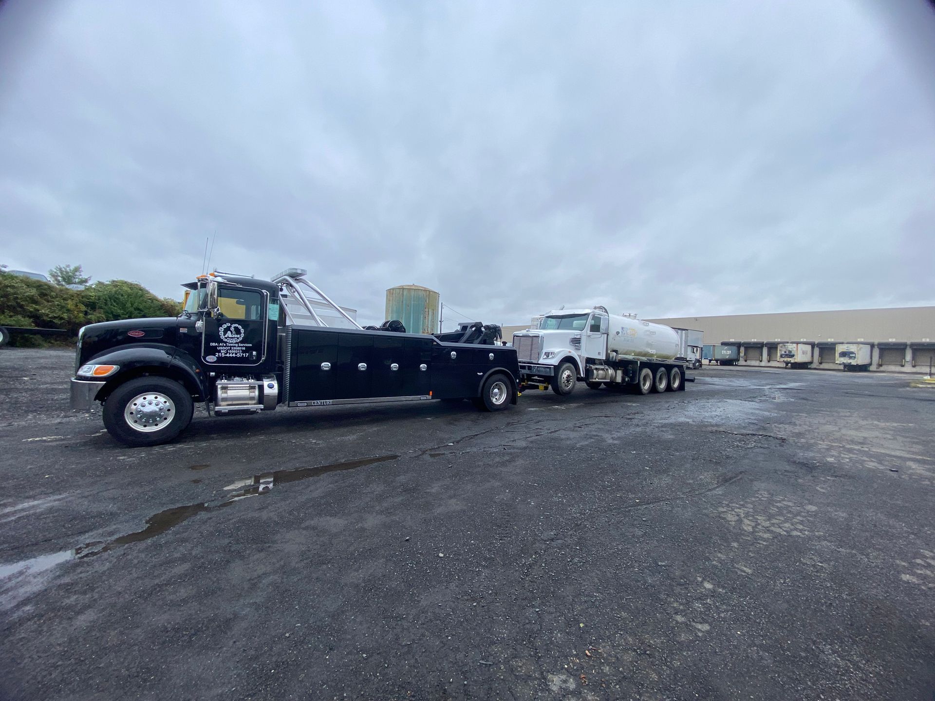 Black tow truck towing a white tanker truck on an asphalt lot under an overcast sky.