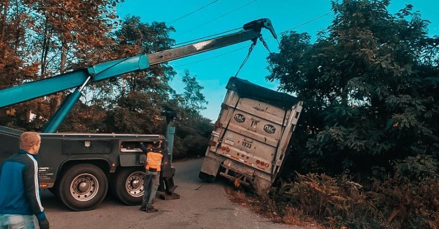 Tow truck lifting a white truck trailer from roadside bushes, with people watching.