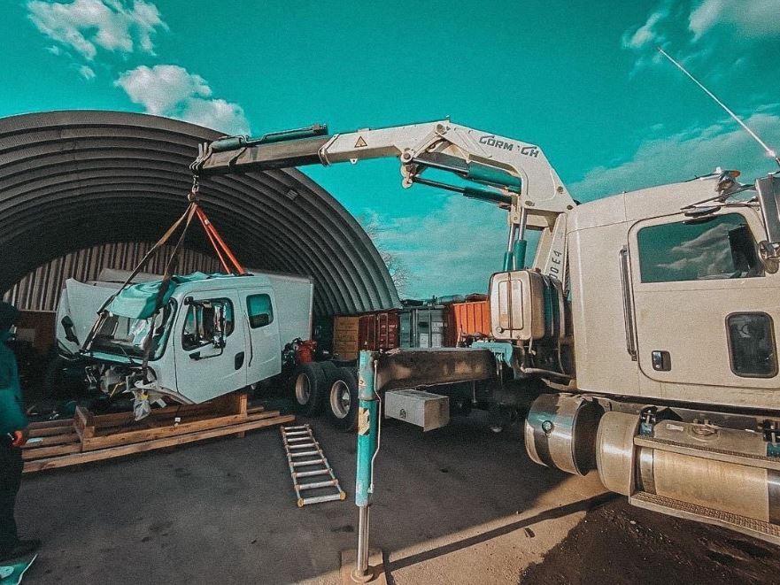 A truck crane lifts a white truck cab from a pallet inside a metal arched structure under a blue sky.