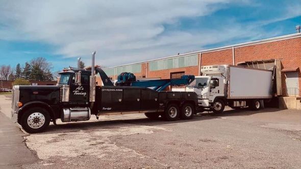 Black tow truck towing a white box truck near a red brick building on a sunny day.