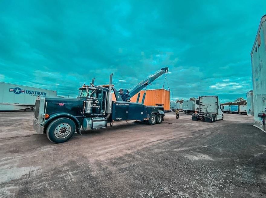 Dark tow truck parked in a lot with a raised boom under a turquoise sky.