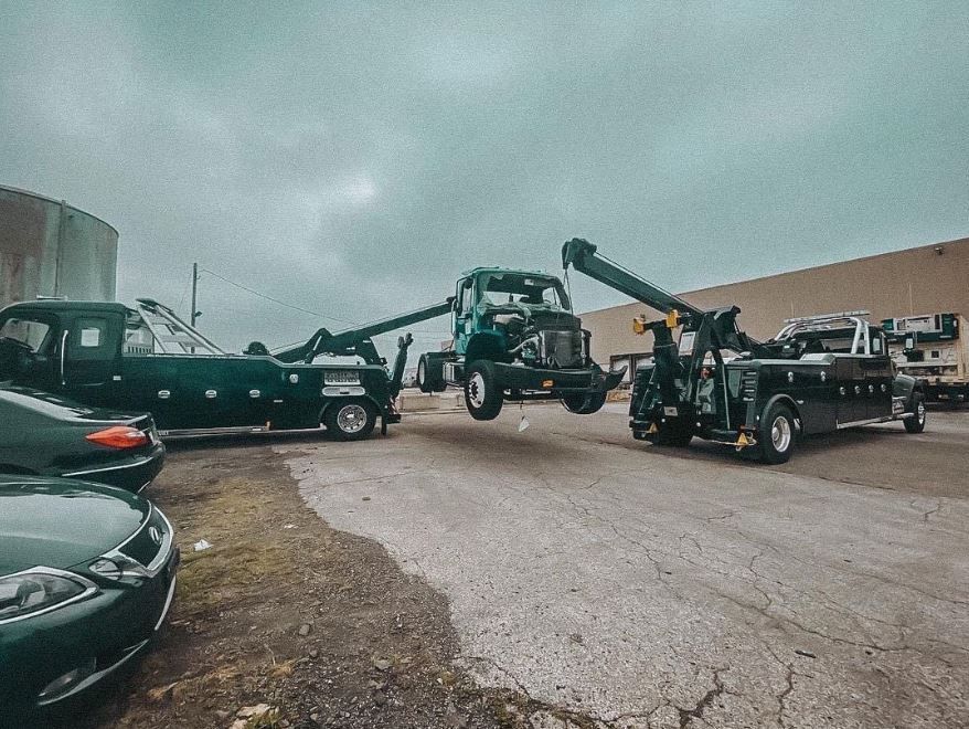 Three tow trucks lifting a green truck on a gray asphalt road, overcast sky.