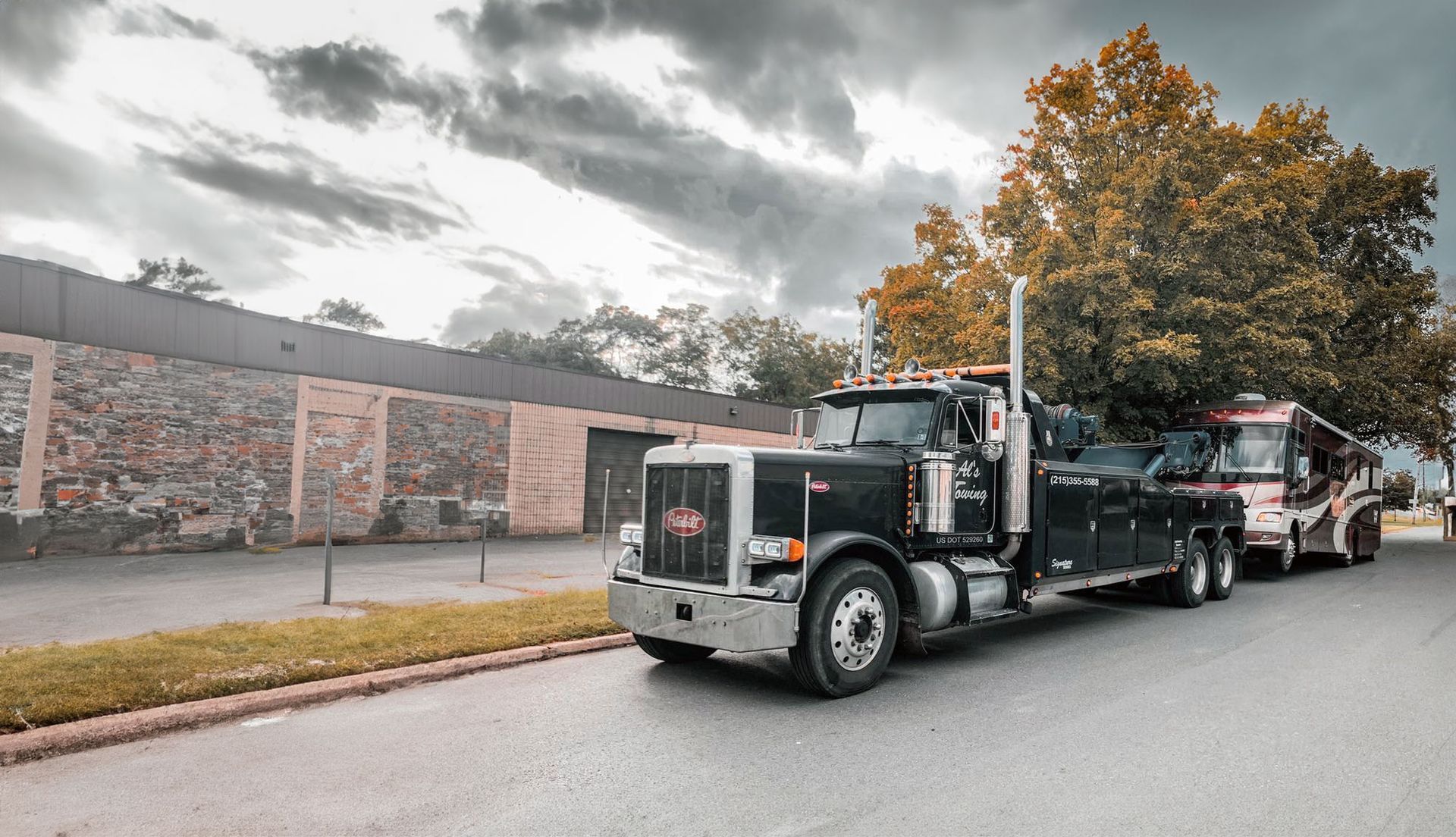 Black tow truck parked on a street, towing a red and white trailer under a cloudy sky.