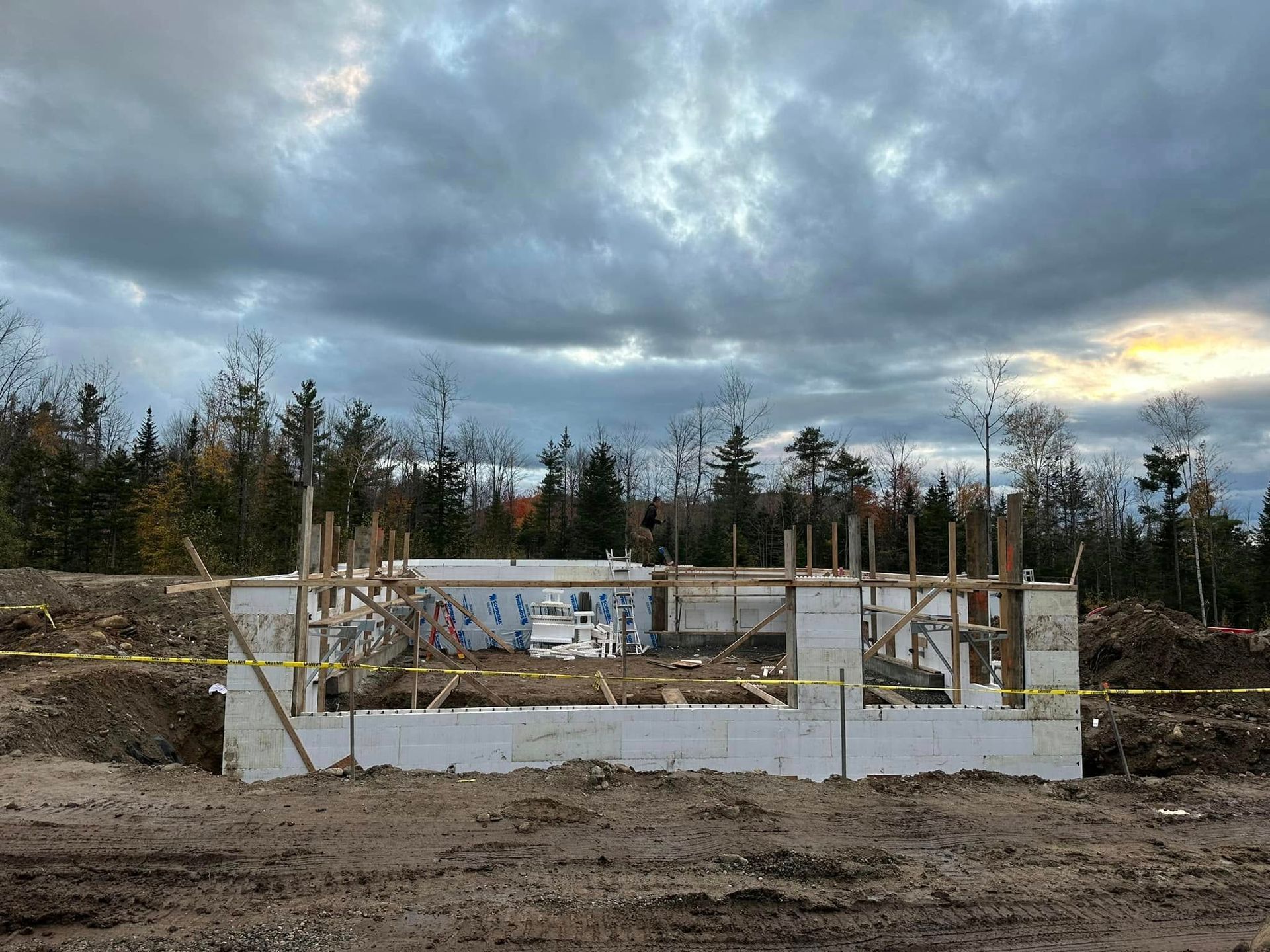 Construction foundation in a muddy clearing under a cloudy sky, with concrete walls and wooden framing.