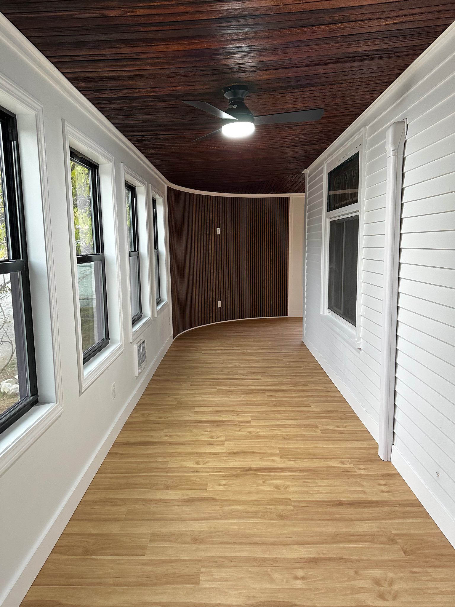 Long narrow enclosed porch with wood floor, white walls, windows on both sides, and a dark wood ceiling.