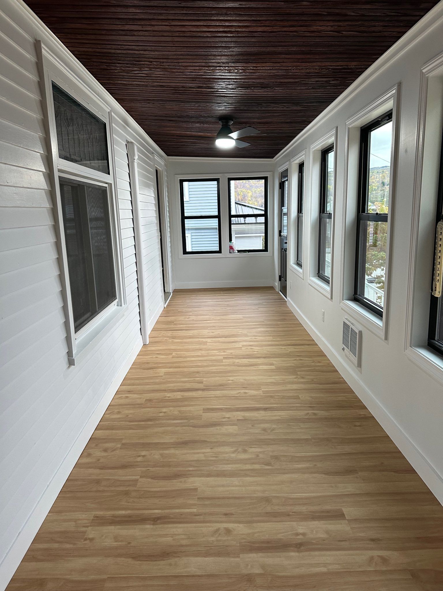 Narrow enclosed sunroom with white walls, wood-look floor, and multiple windows on both sides and end.