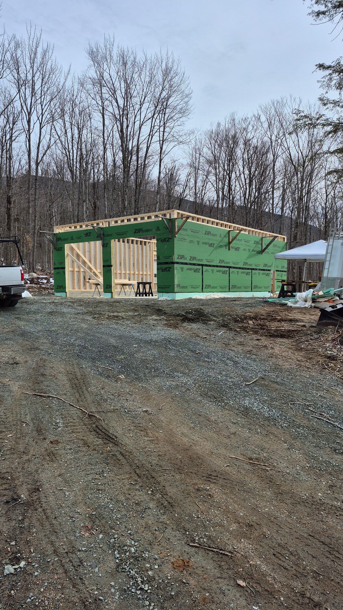 Green house under construction on a gravel lot, with bare trees in the background.