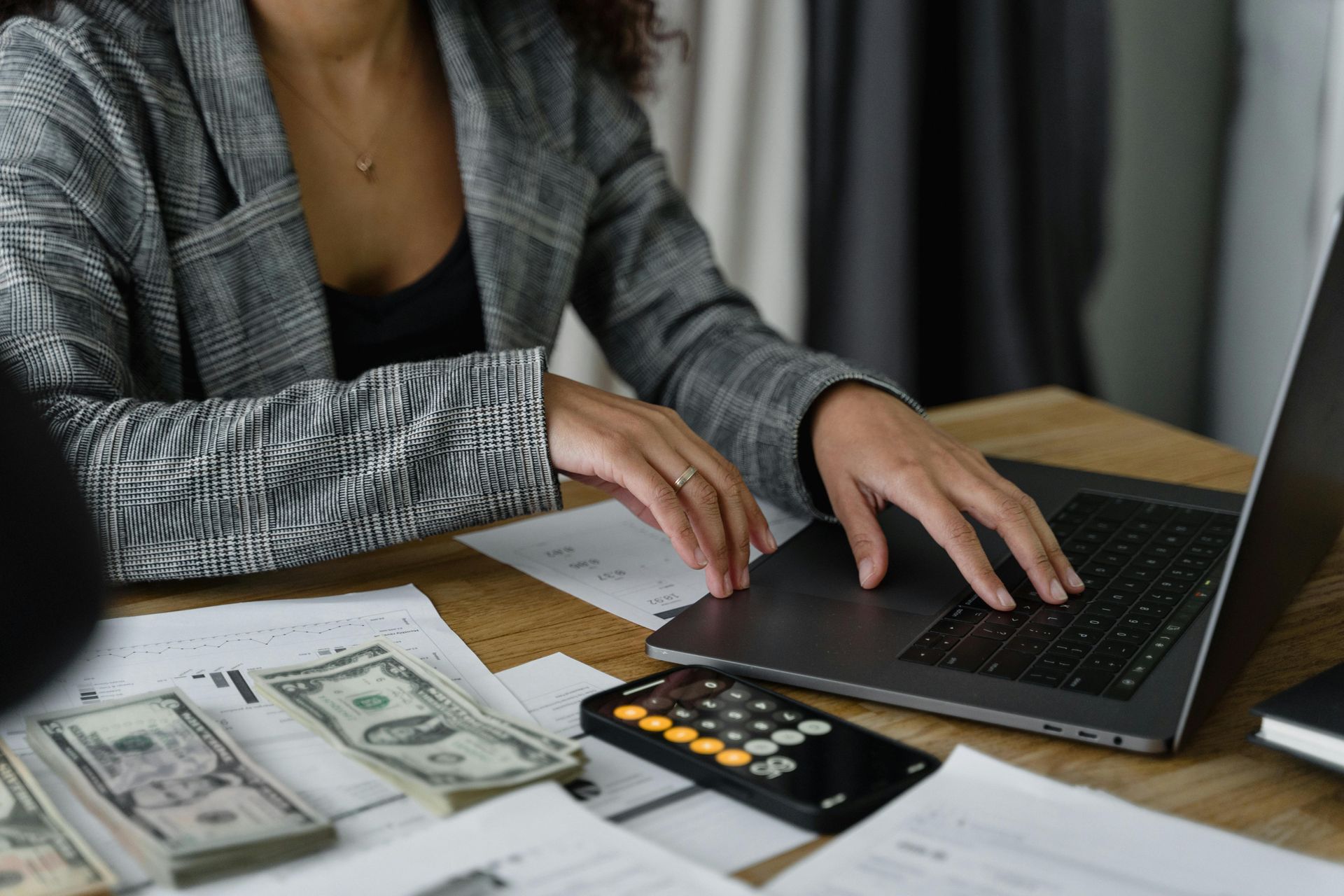 Woman in blazer works on laptop, with money, calculator, and papers on a desk.