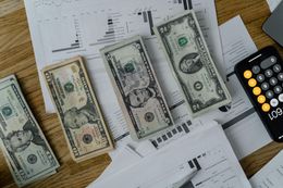 Stacks of US dollar bills and a calculator on papers with graphs.