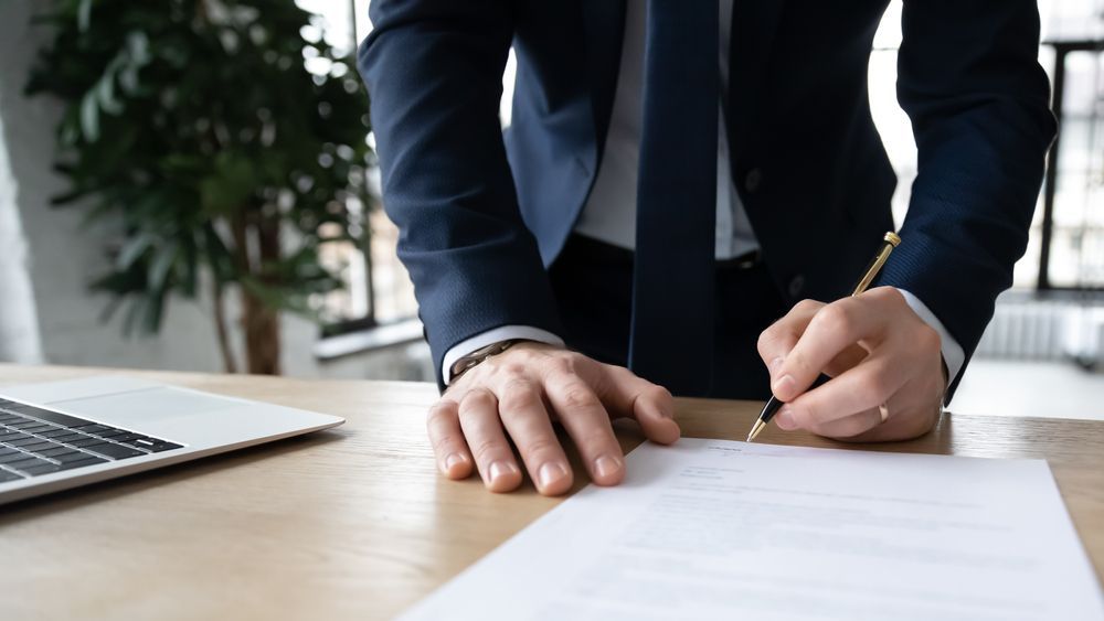 Person in a Navy Suit Signing a Document at a Wooden Desk With a Laptop — Umbrella Legal in Gosford, NSW