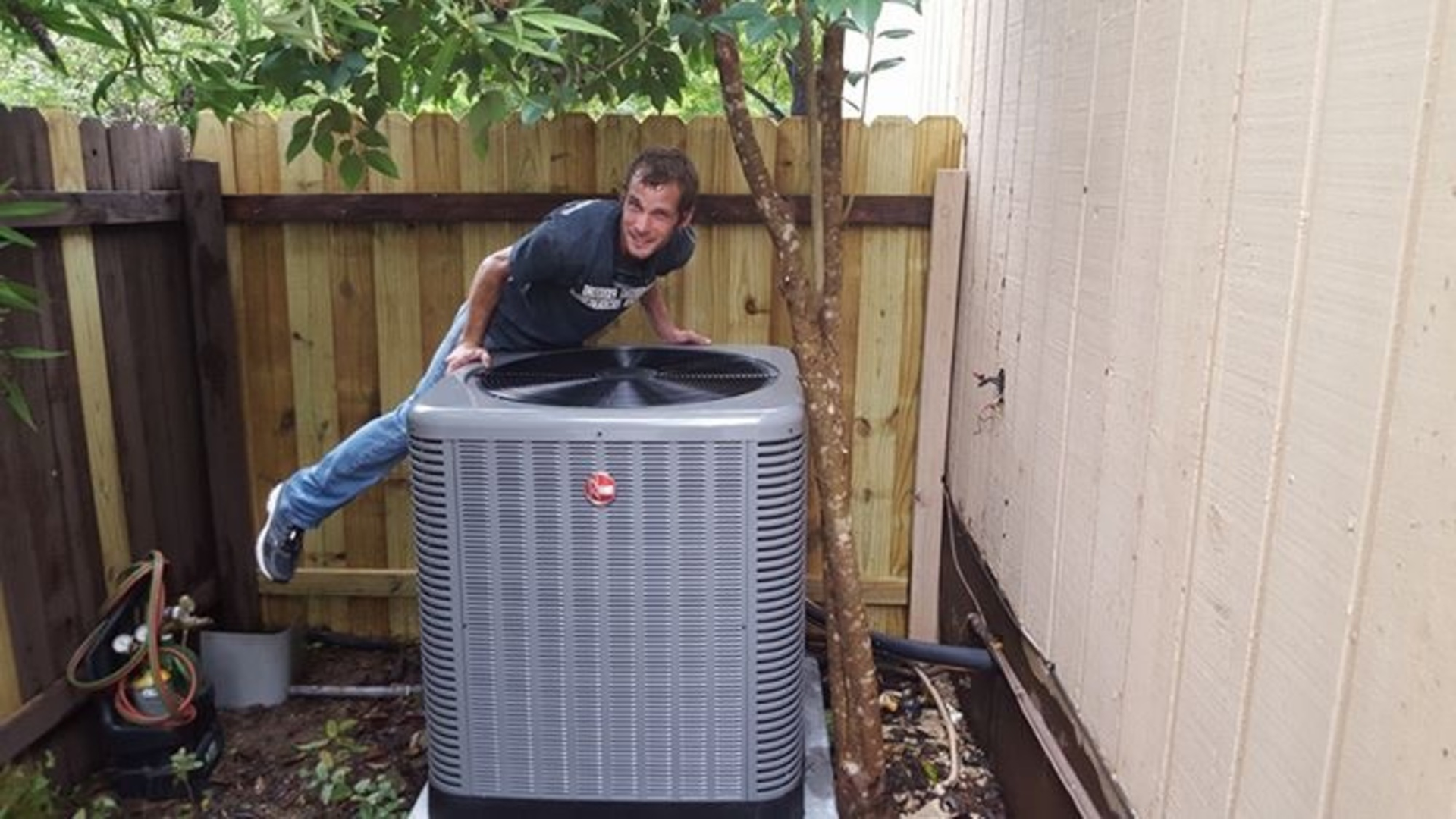 A man is standing next to an air conditioner in a backyard.