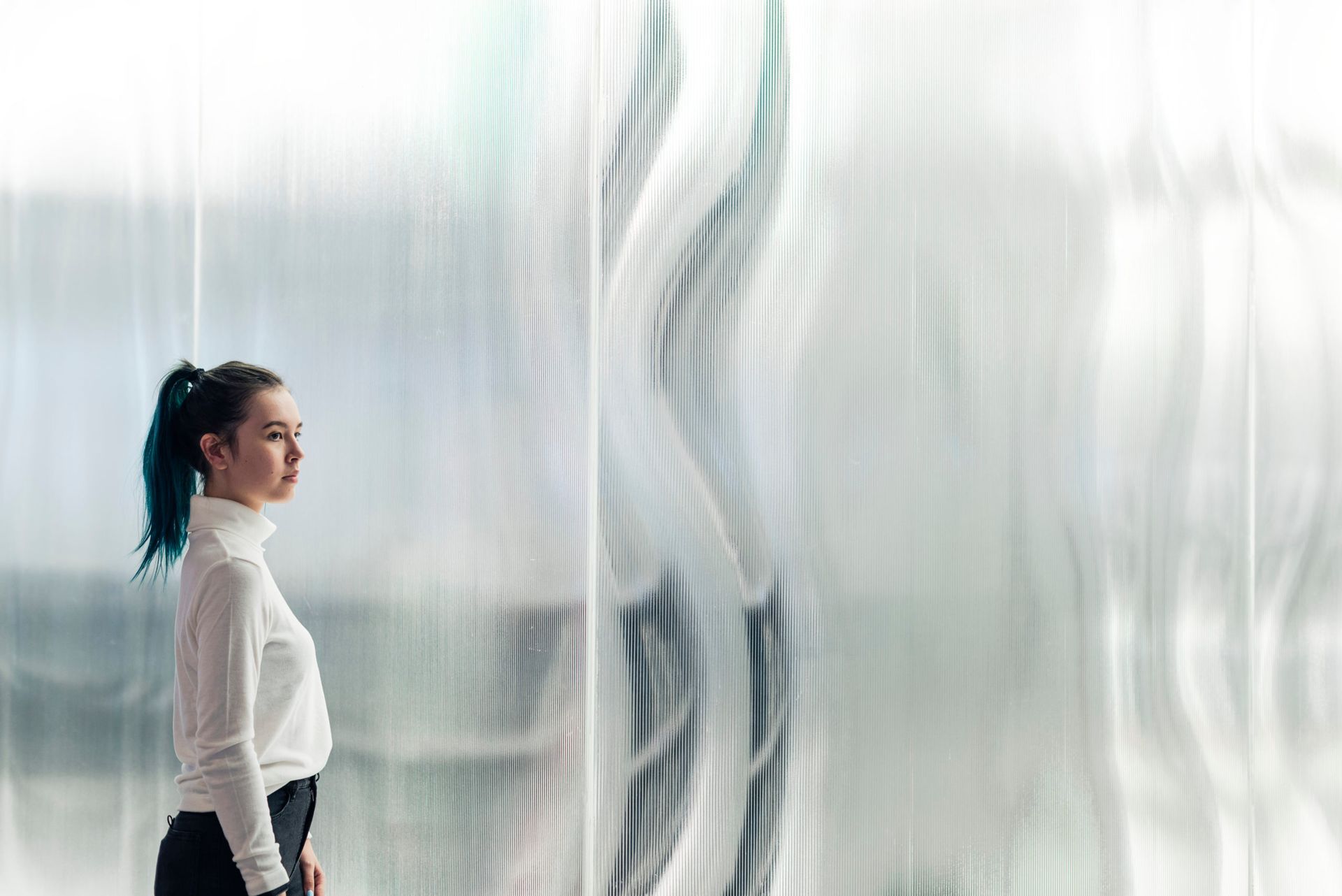 Woman in a white blouse stands beside a rippled translucent wall in a bright, minimalist space