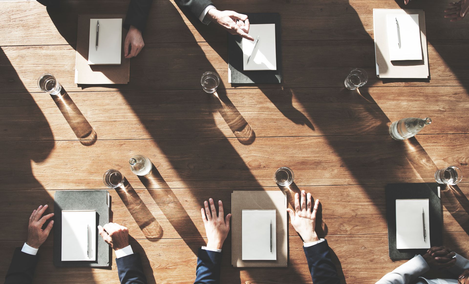 Hands around a sunlit conference table with notebooks, glasses, and coffee, viewed from above.